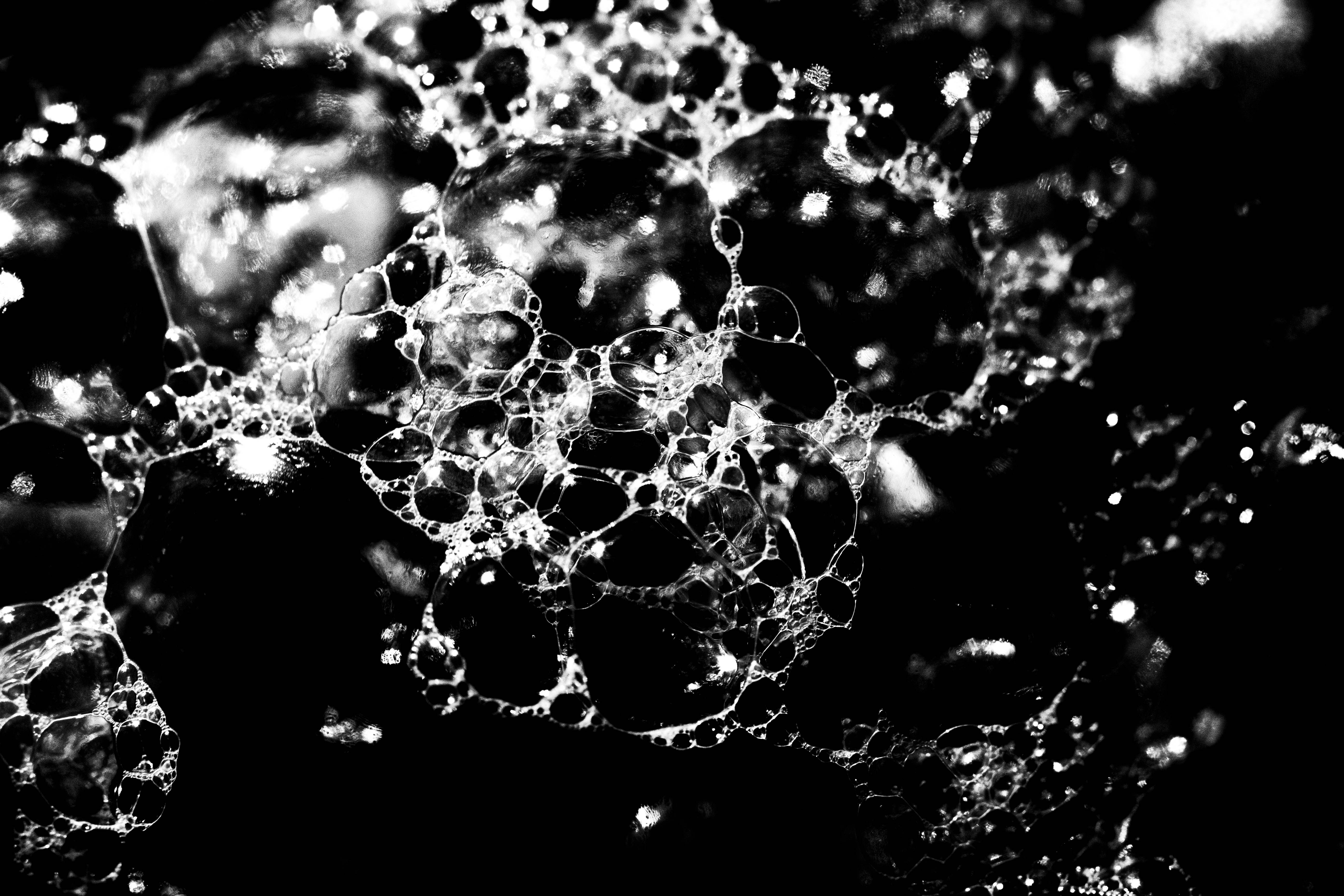 A black and white macro shot of seafoam taken on a digital camera. This image provides a closer look at the bubble-like formations of seafoam. It shows the white outlines of the bubbles enlarged further across the image from the left hand side to the right. In the middle of the bubbles we can see the blackness of the background and some grey-white smudges where the light reflected off the foam, showing off the texture of the bubbles.