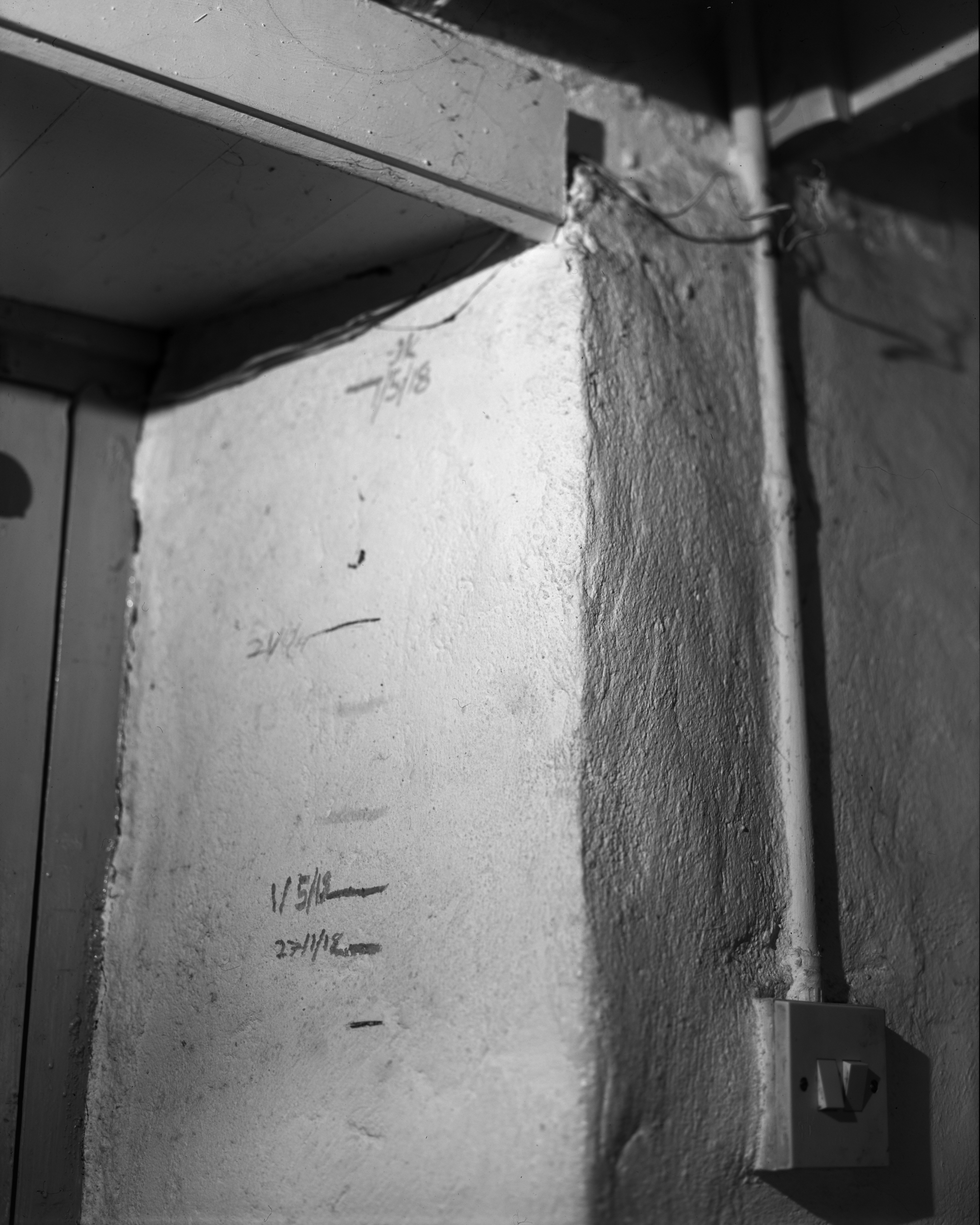 A black and white photograph of a rough, plastered interior wall with several hand-drawn horizontal lines and dates marking different heights, likely tracking a child's growth over time. The wall is next to a doorframe and has an exposed electrical switch and conduit running vertically. Some of the markings are labeled with dates, such as "1/5/18" and "27/11/12." The scene is softly lit, highlighting the wall’s texture and the personal, lived-in feeling of the space.