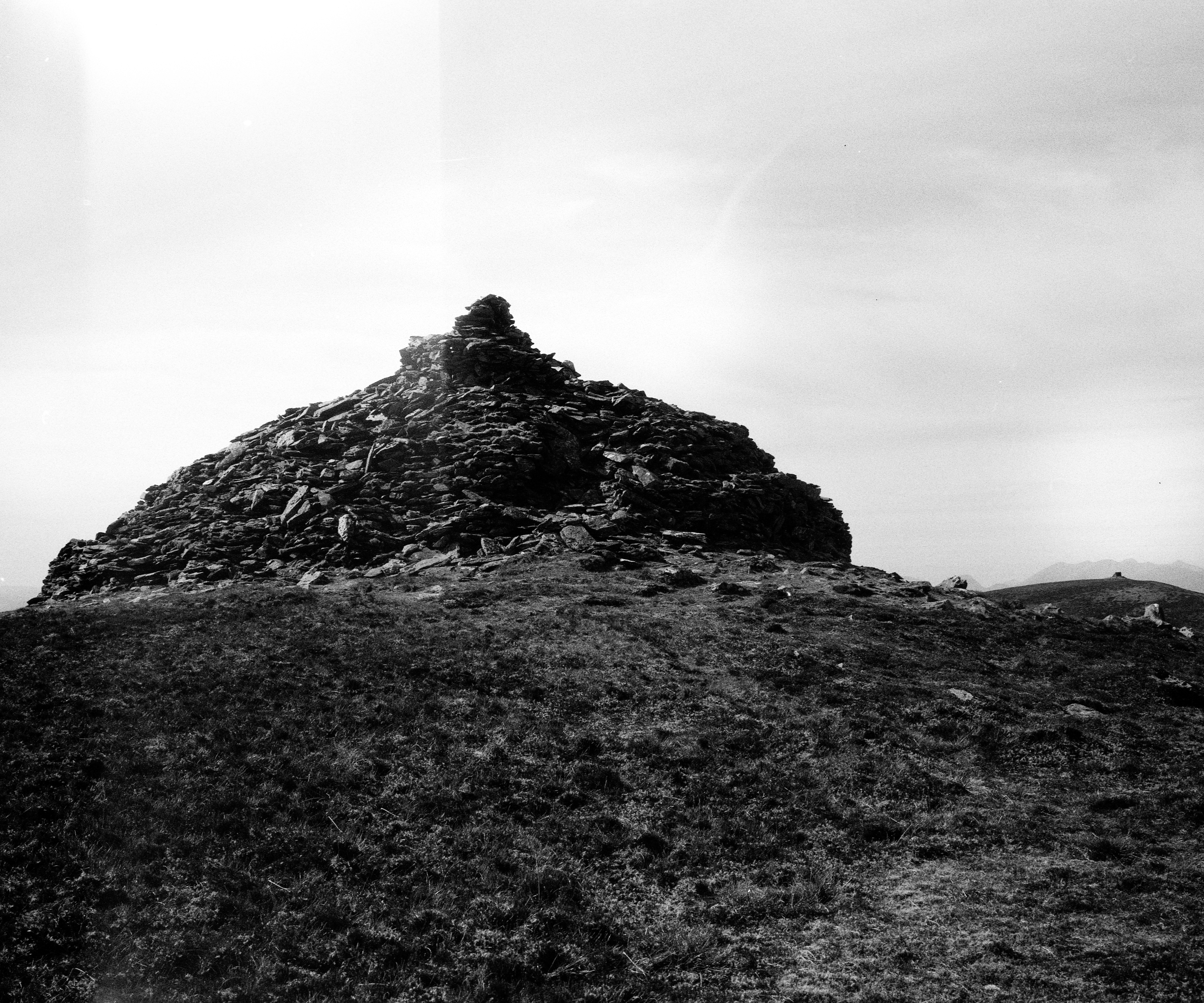 Black and white photograph of a large cairn made of stacked stones, situated on a grassy mountaintop. The cairn rises in a conical shape, its dark texture contrasting against the bright, overcast sky. Distant parallel hill of mountain peak with similar structure is visible on the horizon, contributing to the importance of the location with a clear feeling isolation and elevation.
