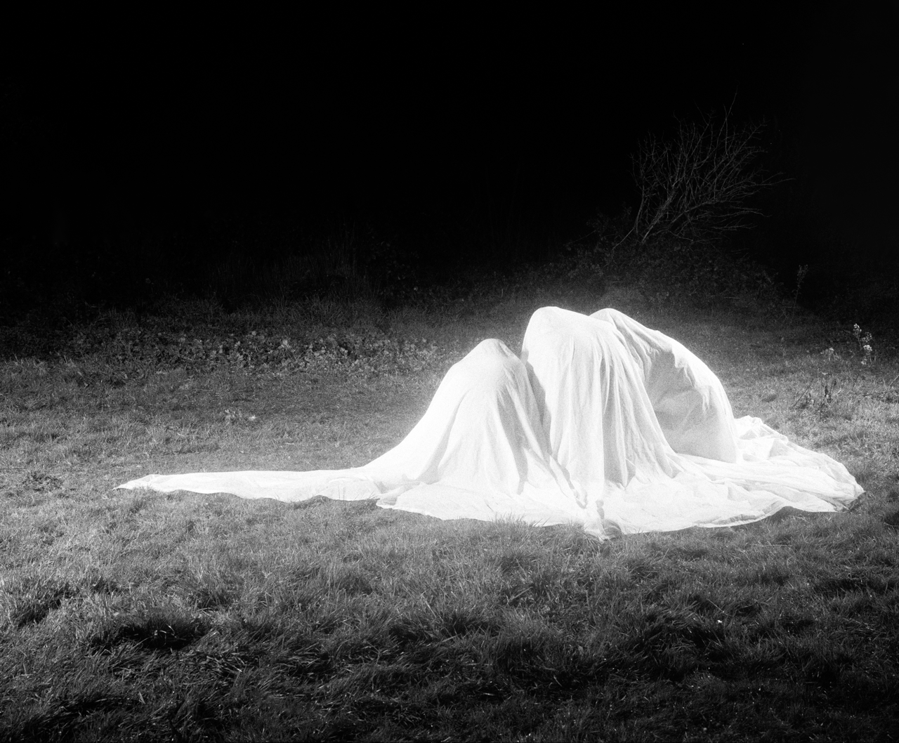 A black and white photograph depicting three human-like forms covered in a large white sheet,  kneeling on a grassy field at night. The scene is dramatically lit, casting a glow on the sheet and surrounding grass while the background fades into darkness. Sparse vegetation and a bare shrub are visible at the edge of the illuminated area, adding to the eerie and surreal atmosphere