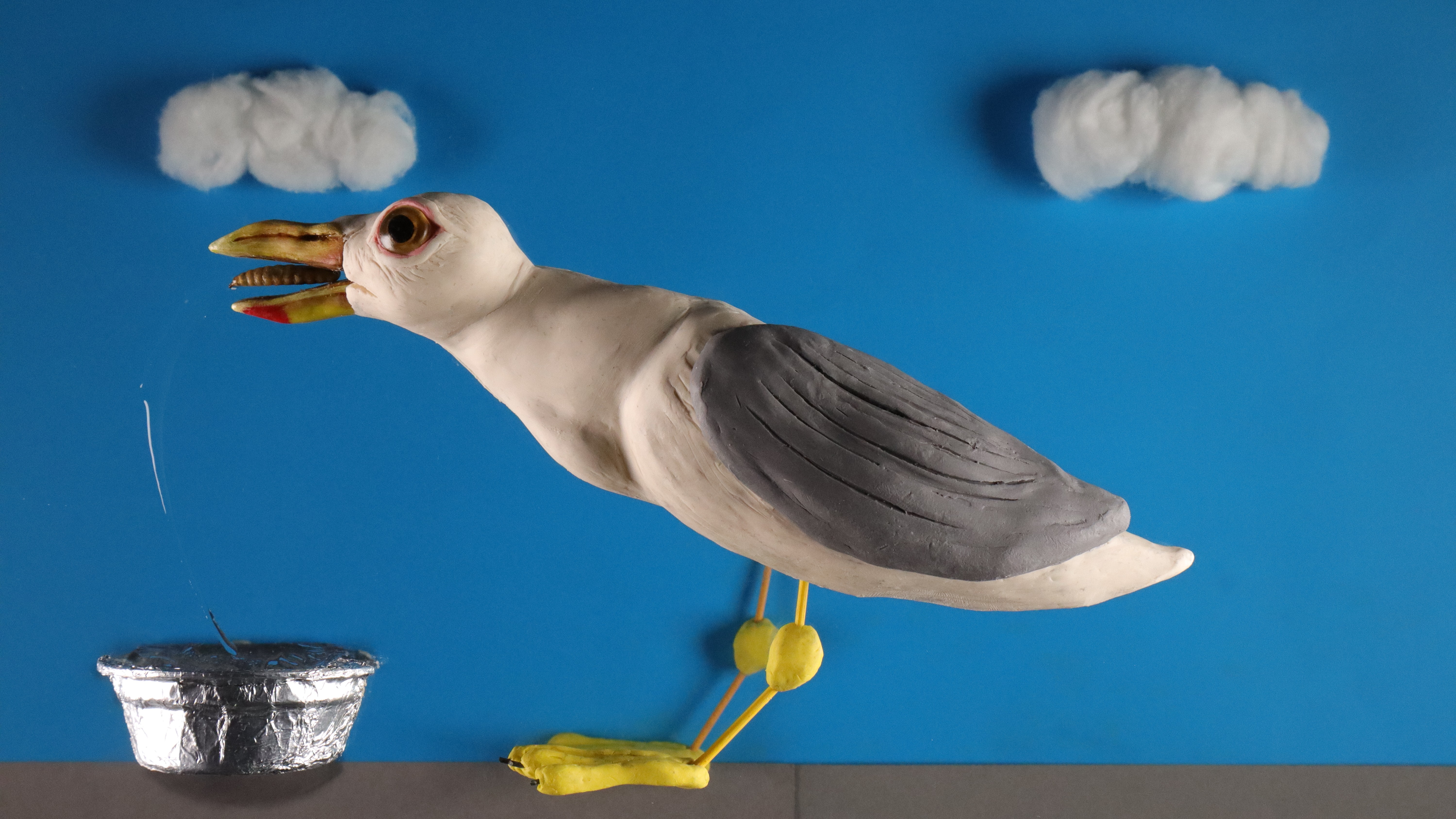 A still of a claymation scene on a multiplane. A plasticine seagull is mid chomp on a dried mealworm from a tinfoil dog bowl, with a trail of hair gel representing a water splash. 
