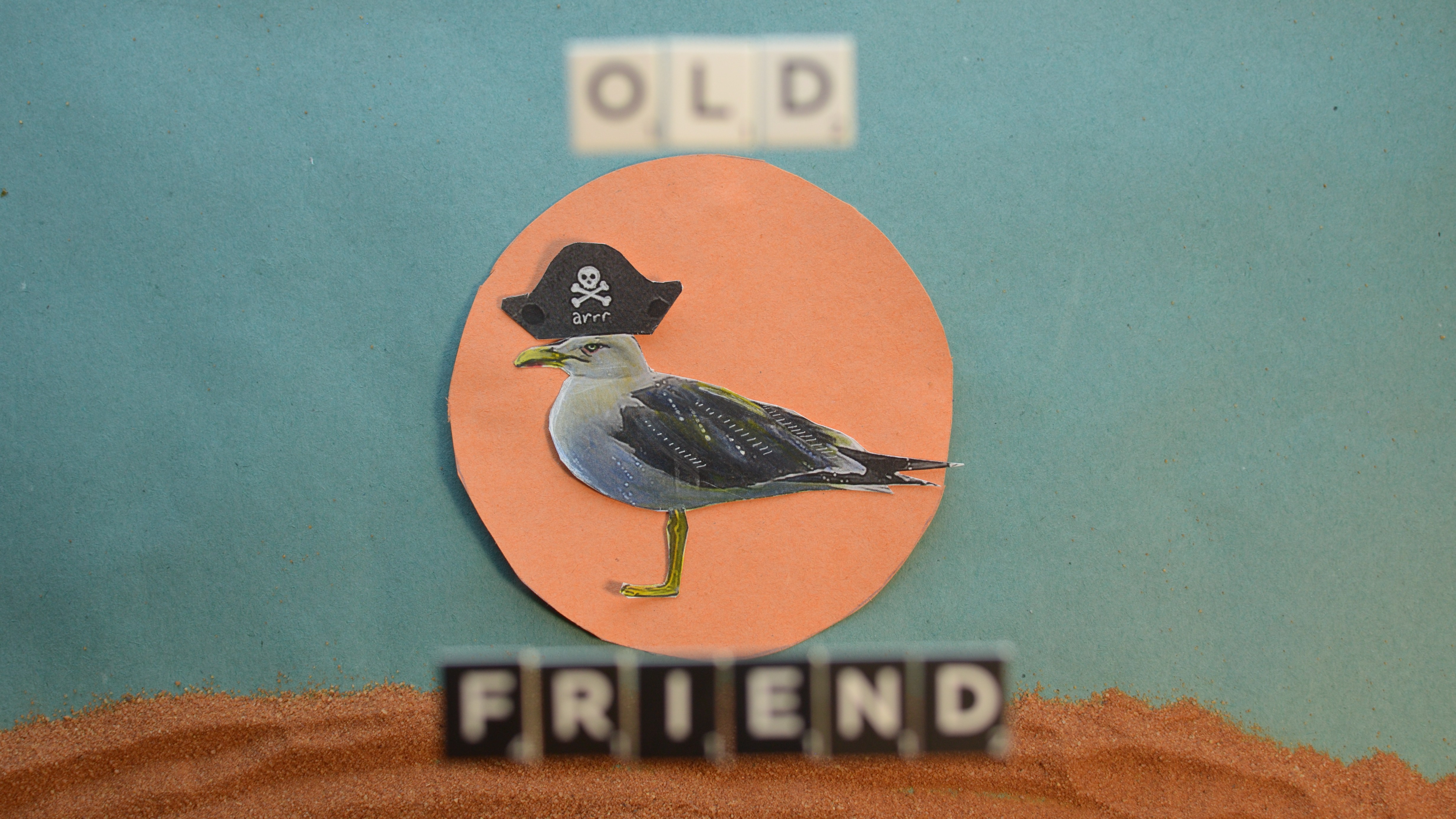 A printed cutout image of a seagull wearing a pirate hat standing in an orange paper circle on a blue sugar paper background, with red sand underneath. Out of focus scrabble tiles in the foreground read "OLD FRIEND"