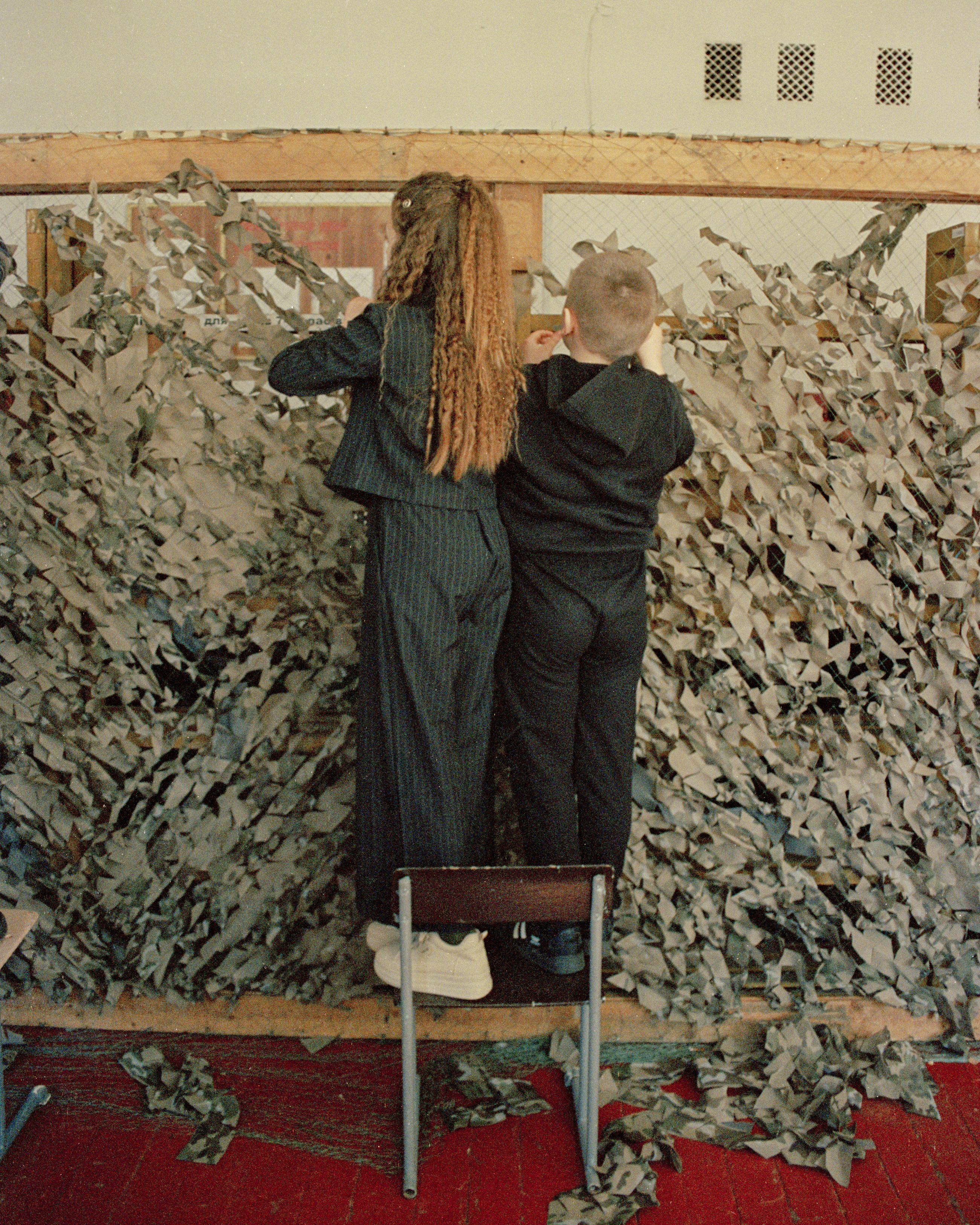 Two kids stand on the chair making a camouflage net turned away from us