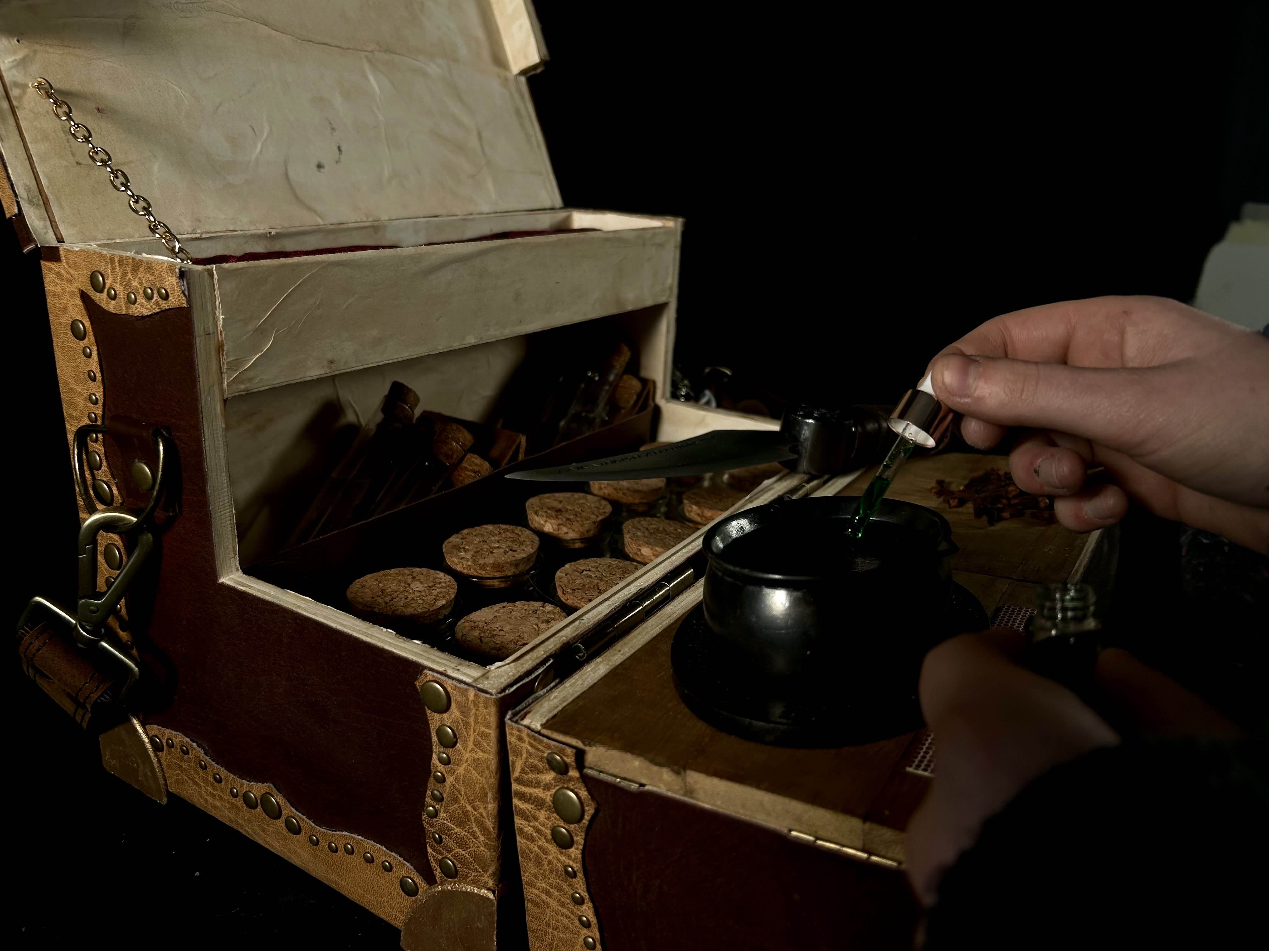 Close up of the open box. There are cloves scttered on the cutting board with the knife resting next to it. A hand is dropping in green liquid into the cauldron, taken from the left side.