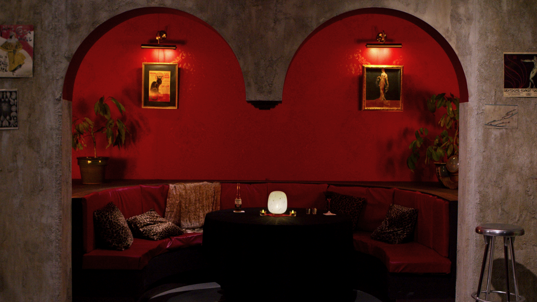 Photo of an alcove framed by two arches. The arches are grey cement, while the alcove walls are red. There is a red couch and a black cloth covered table in the alcove.