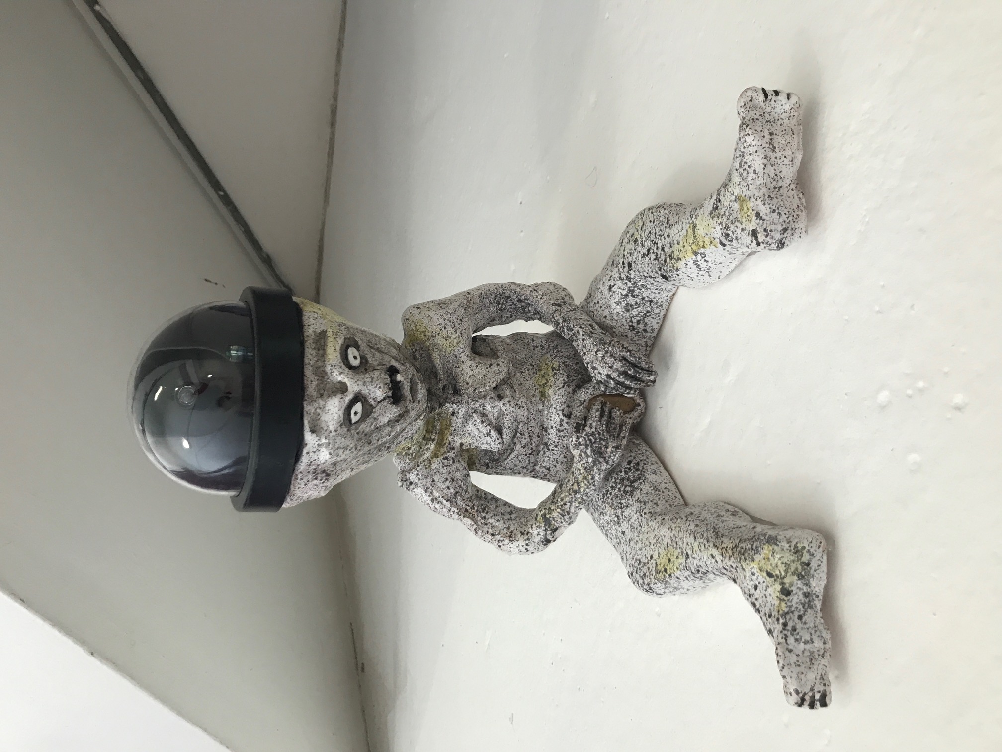  Image of a ceramic Sheela-na-gig, in a sitting position with legs outstretched wearing a dome CCTV camera on her head like a hat.