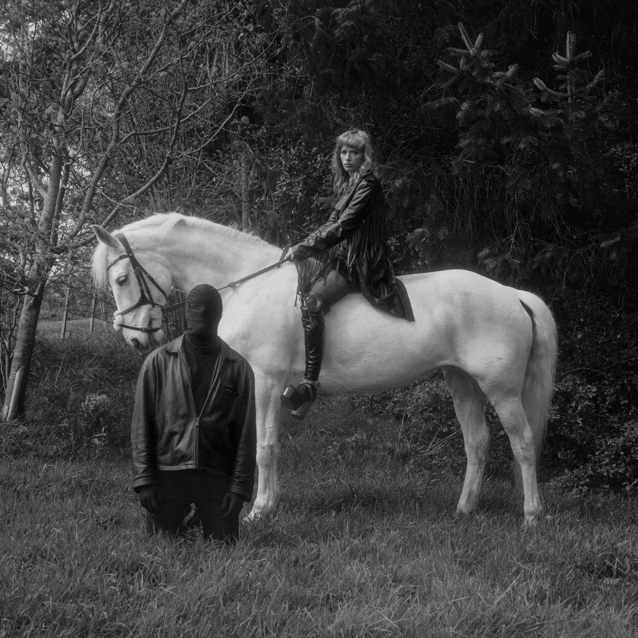 This image is a medium format photograph in black and white depicting a Femdom, her horse and her submissive. The Femdom is on top of the horse while the submissive kneels beside them with their hands by their sides. They are in a grassy area surrounded by trees.