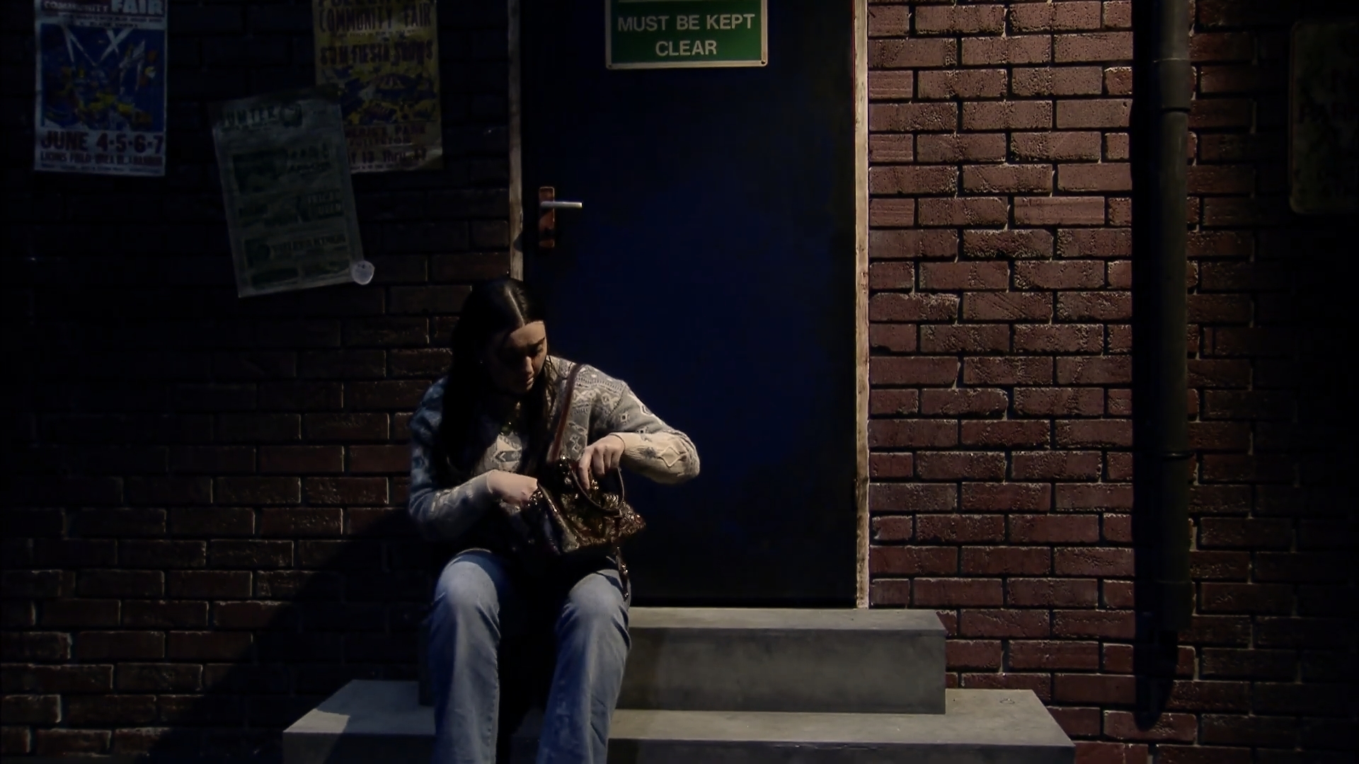 Photo of a woman sitting on steps in front of a brick wall with a blue door. It is dark except for light from a street lamp.