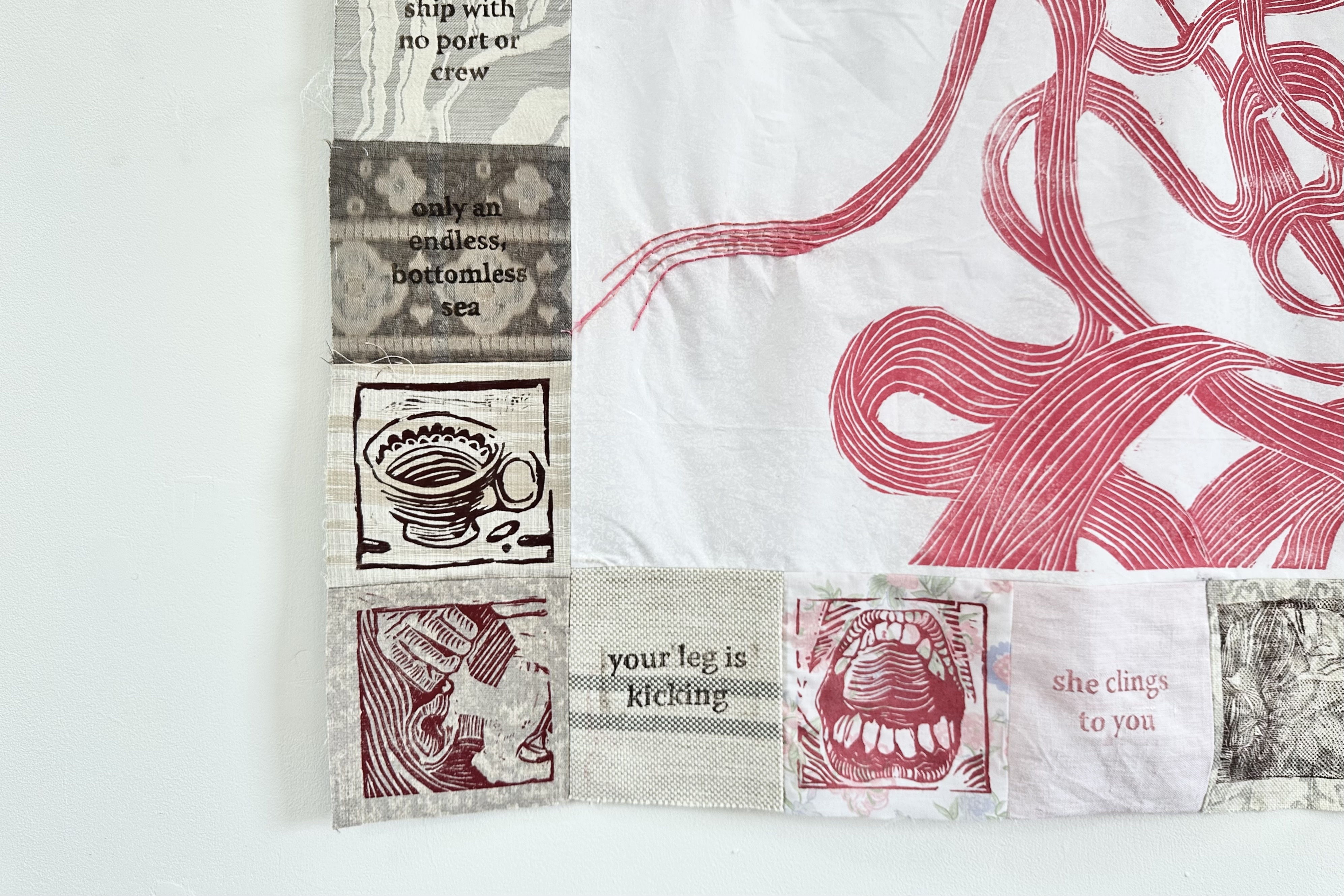 A closeup of the quilt. There is embroidery visible continuing off of the pink lines in the centre, and a border of scrap fabrics. A print of a cup, a face and a mouth, with text in the squares between and around them.