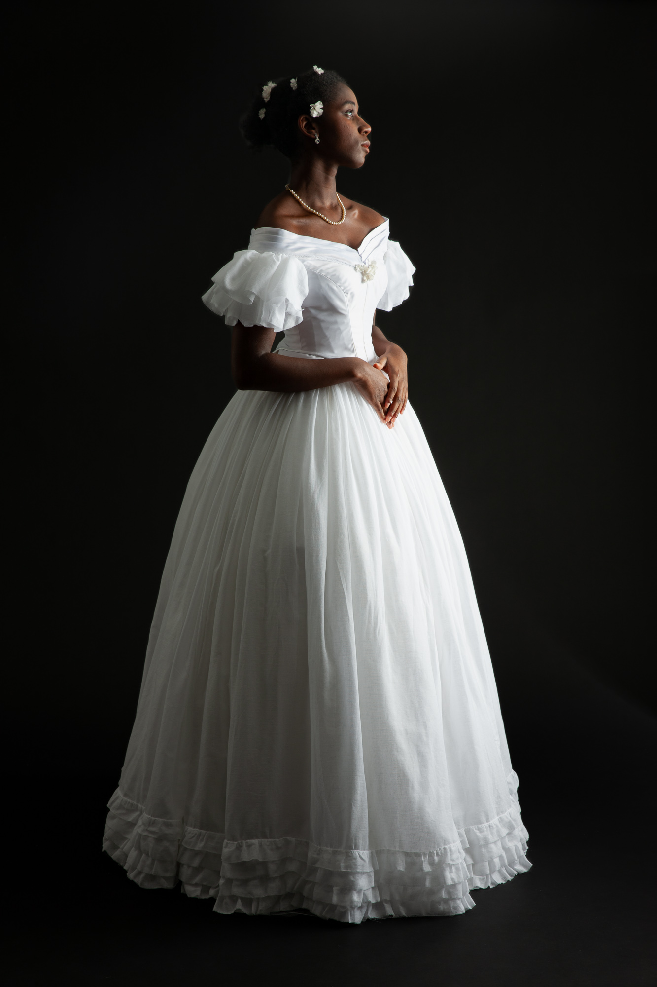 A woman in a full-skirted white dress with short, ruffled sleeves and white flowers in her hair standing in profile.