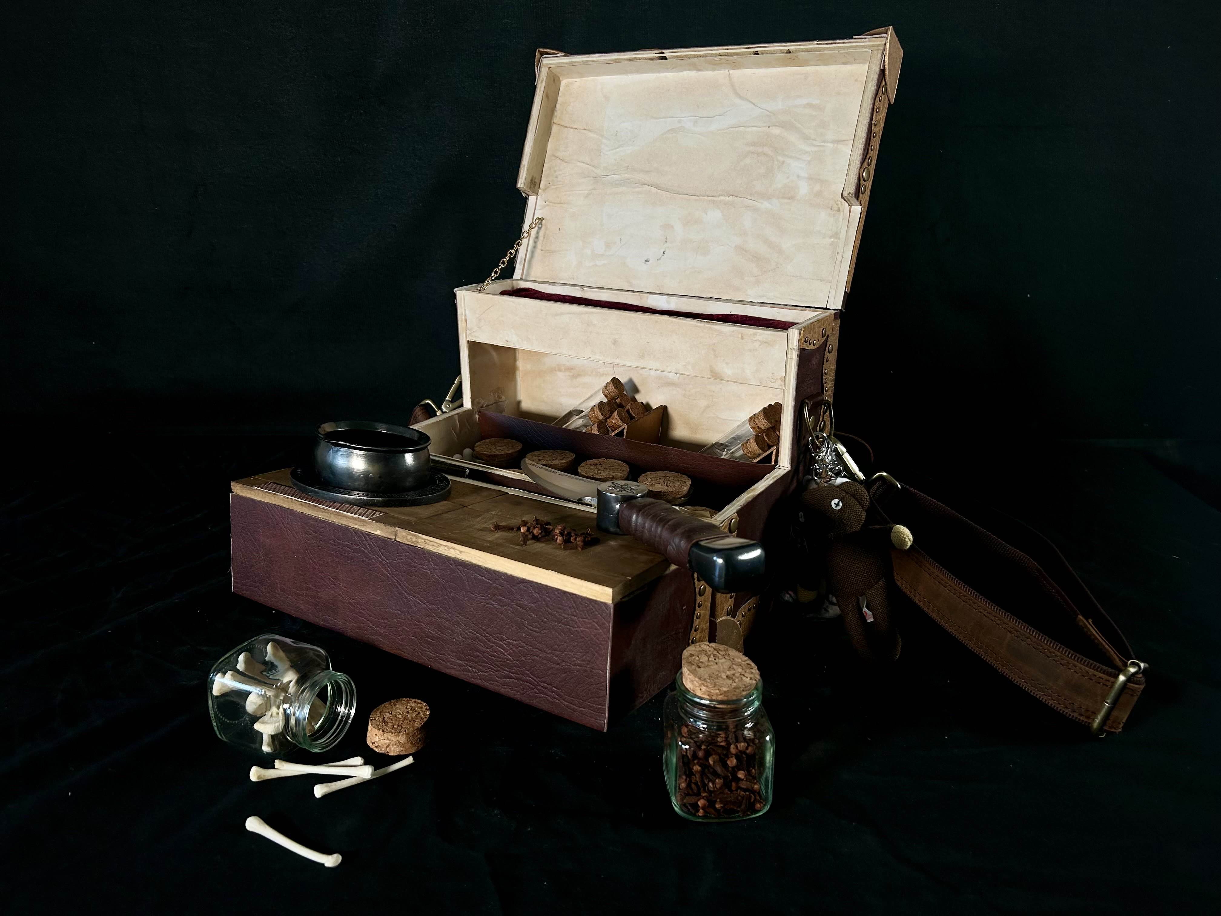 an open box containing many jars and test tubes, with the top folded out to hold a cauldron and a chopping board with cloves scattered on it and a knife on top. In front is a tipped over jar or small bones and an upright jar of cloves 