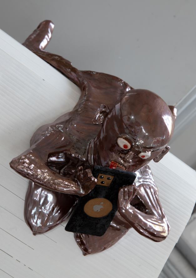 Image of Copper colored Sheela-na-gig lying prone over a small bump holding an iphone and attempting to take a video
