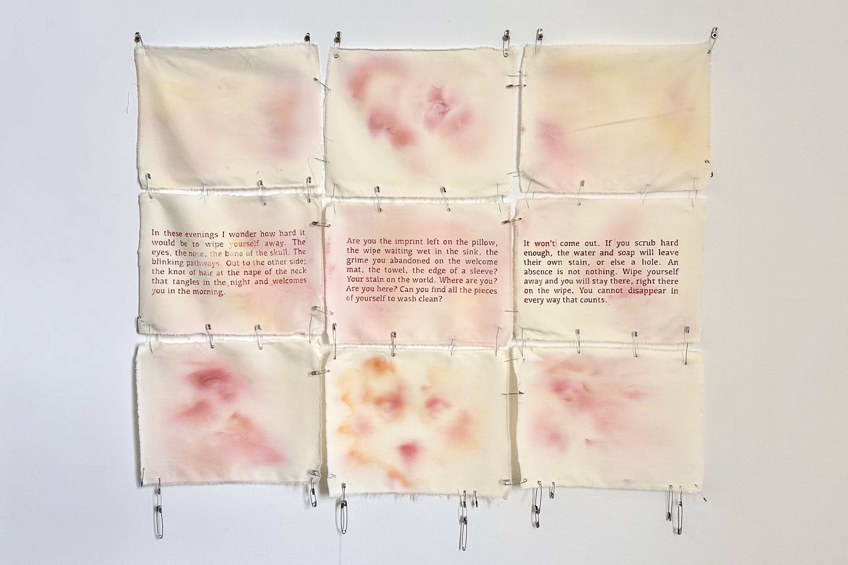 A 3x3 grid of yellowed rectangular pieces of cloth held together by safety pins. In the middle row, there’s printed text on each one, while in the other rows, there are pink and yellow watercolour splotches that resemble faces and eyes.