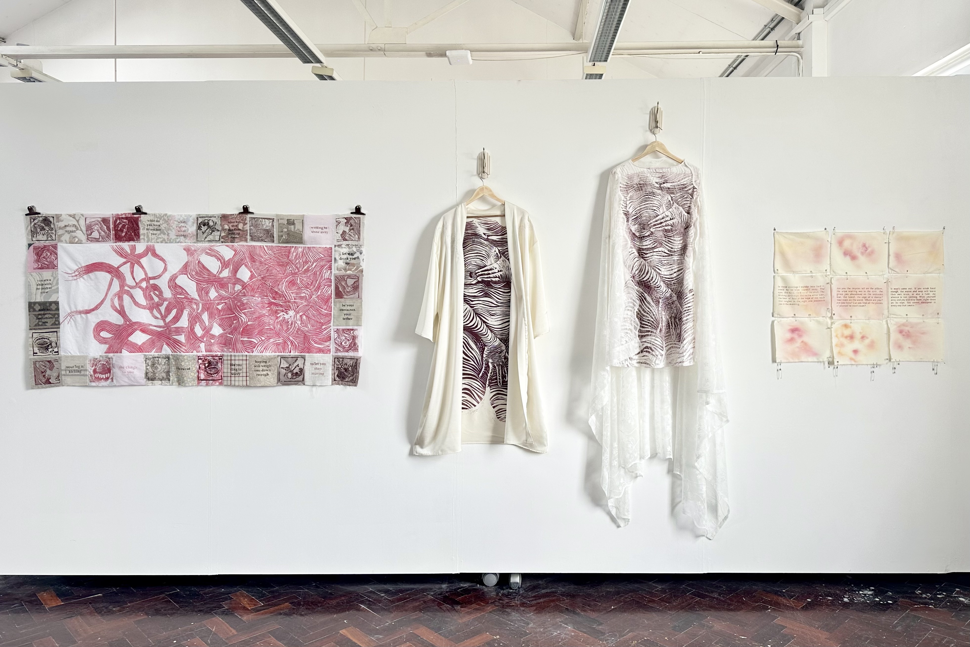 A wide view of the wall. From right to left, there is a colourful tapestry piece with prints on it, a light robe with a woman’s body printed on it, a textured white dress with the same print and hanging lace, then yellowed pieces of cloth with pink images and text connected by safety pins in a grid.