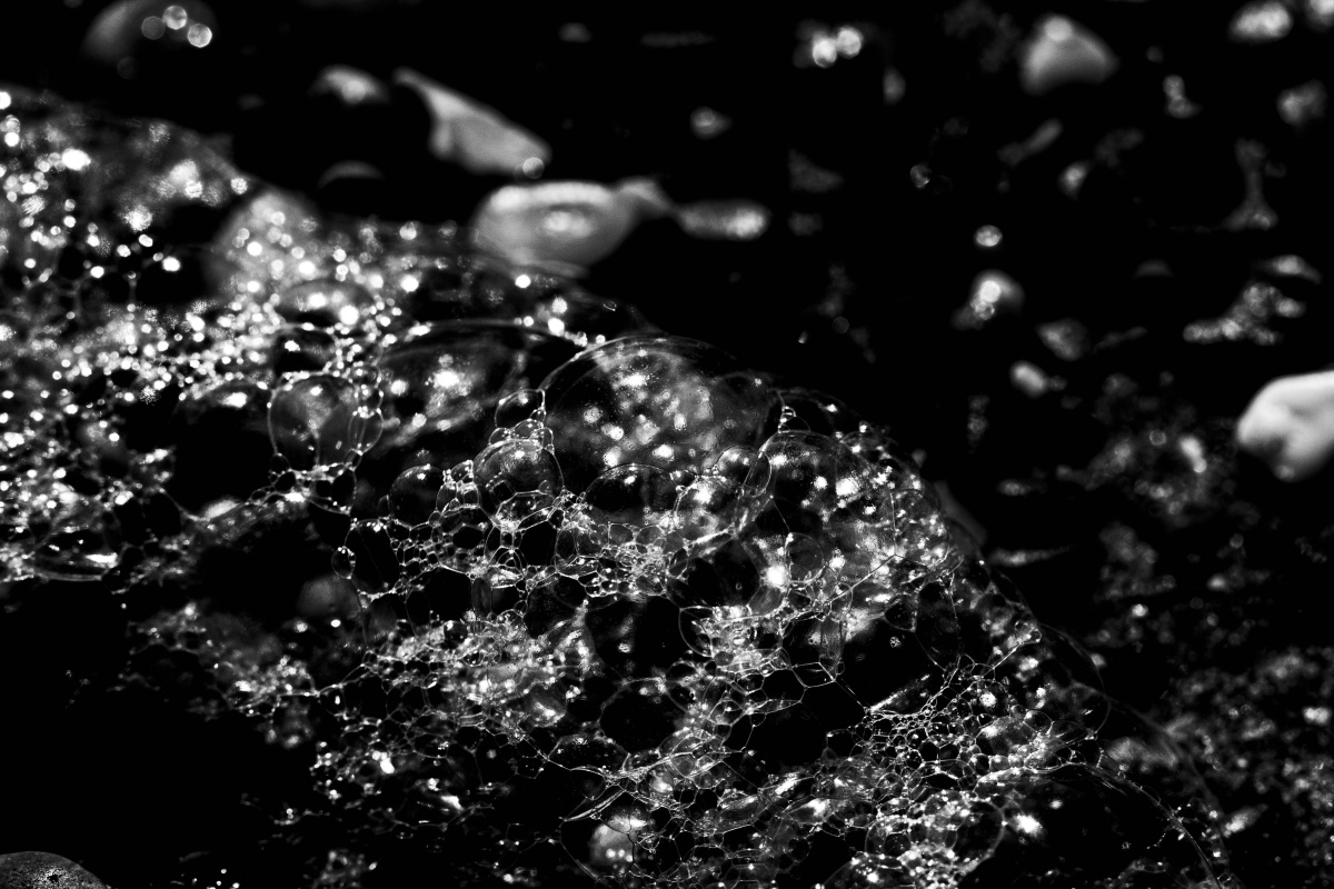 Black and white image taken on a digital camera depicting seafoam on a beach. The bubbly outline of the foam starts on the left hand side of the photo and tapers off in the right bottom foreground. The grittiness of the sandy beach can also be seen in background and right side of the photograph.