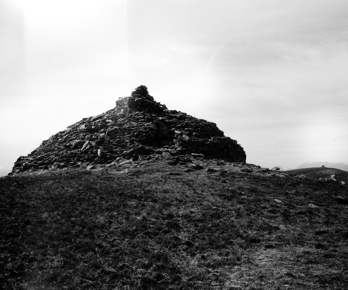 Black and white photograph of a large cairn made of stacked stones, situated on a grassy mountaintop. The cairn rises in a conical shape, its dark texture contrasting against the bright, overcast sky. Distant parallel hill of mountain peak with similar structure is visible on the horizon, contributing to the importance of the location with a clear feeling isolation and elevation.