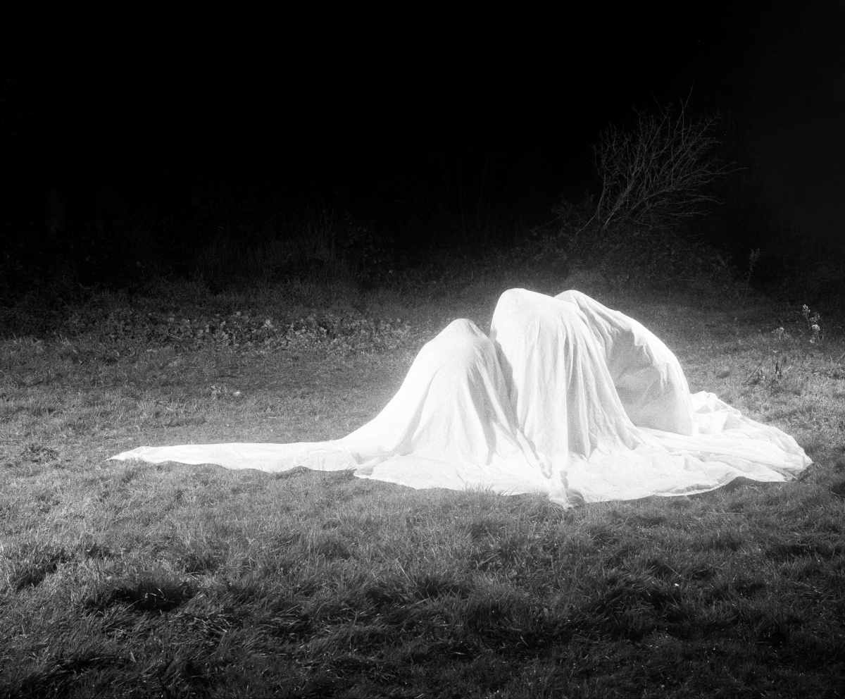 A black and white photograph depicting three human-like forms covered in a large white sheet, kneeling on a grassy field at night. The scene is dramatically lit, casting a glow on the sheet and surrounding grass while the background fades into darkness. Sparse vegetation and a bare shrub are visible at the edge of the illuminated area, adding to the eerie and surreal atmosphere.