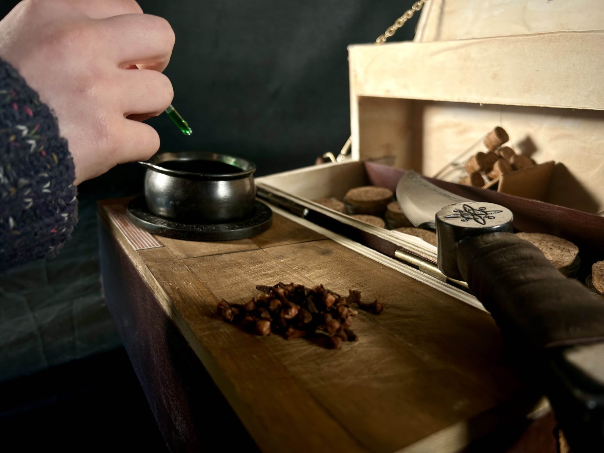 Close up of the open box. There are cloves scttered on the cutting board with the knife resting next to it. A hand is dropping in green liquid into the cauldron, taken from the right-side.