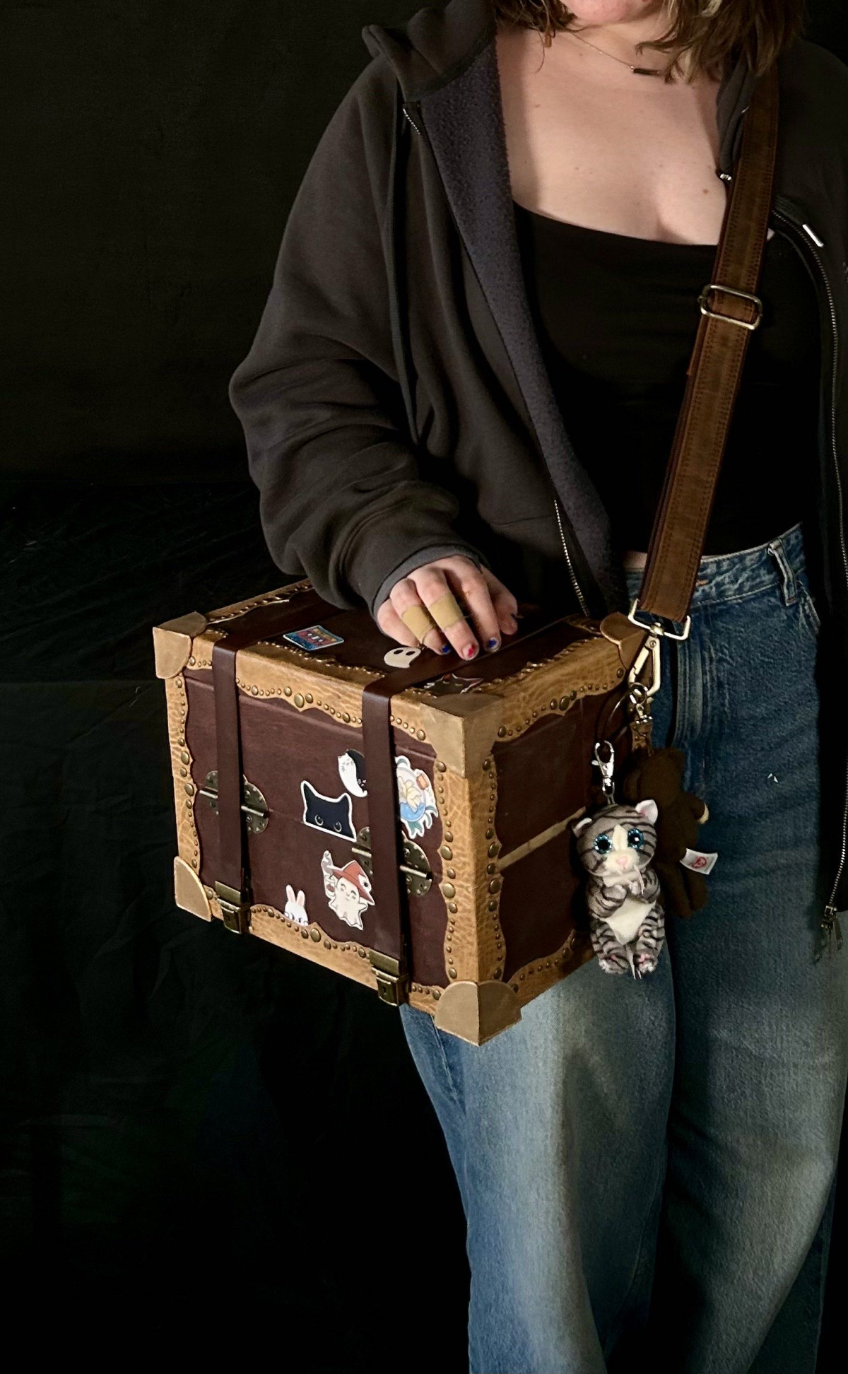 Closed leather-bound box, being worn by the shoulder-strap by someone in jeans and a hoodie. There are stickers on the front and top of cats, ghosts, potion bottles and a rabbit.