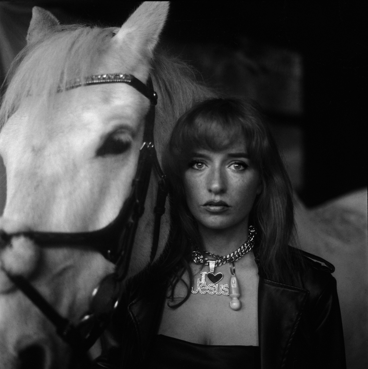 This black and white medium format photograph is a portrait on the Femdom and her horse. The Femdom is in focus in the midground and the horse is out of focus in the foreground. She is wearing a black leather jacket and jewellery on her neck. One necklace says “I love Jesus” and the other is a miniature vibrator.