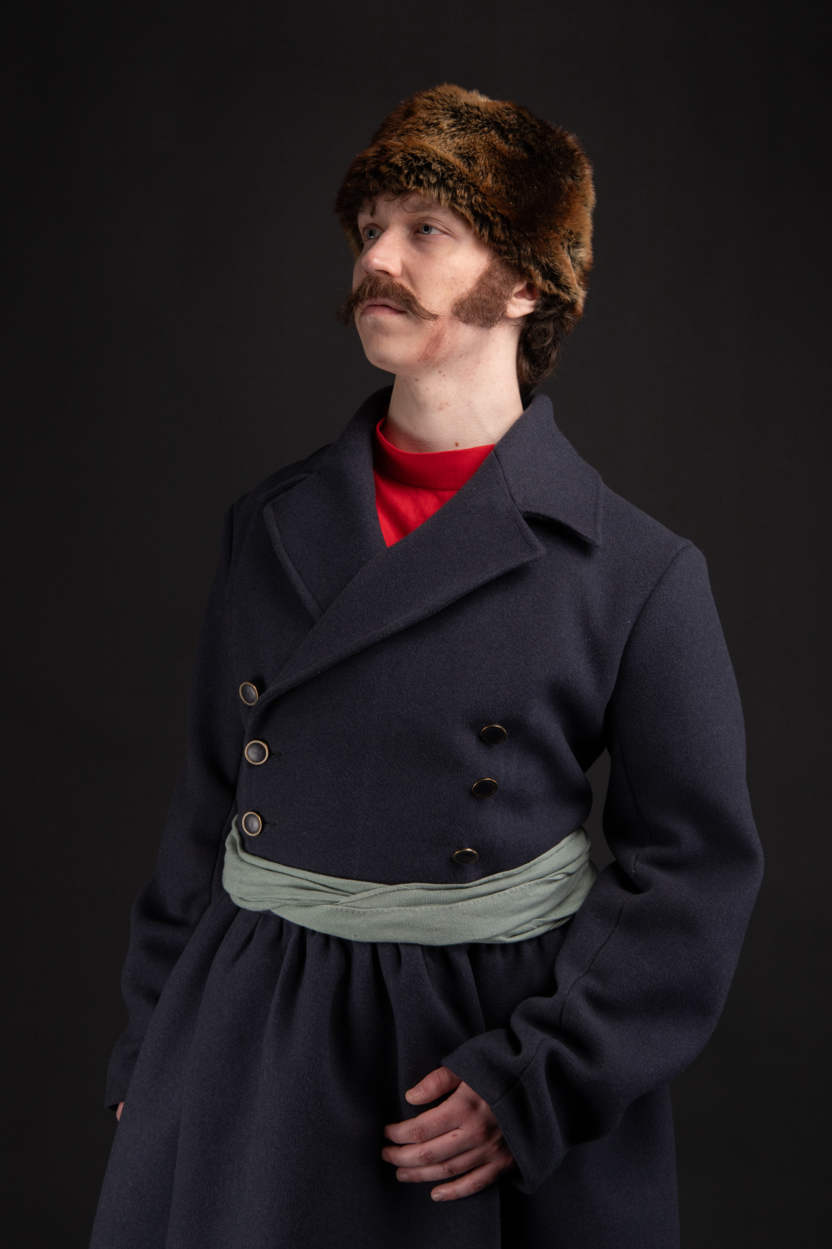 A man with sideburns and moustache wearing a brown fur hat and an army blue, double-breasted, wool frock coat with a sage green sash belt over a red kosovorotka peasant shirt standing at a three-quarter angle. 