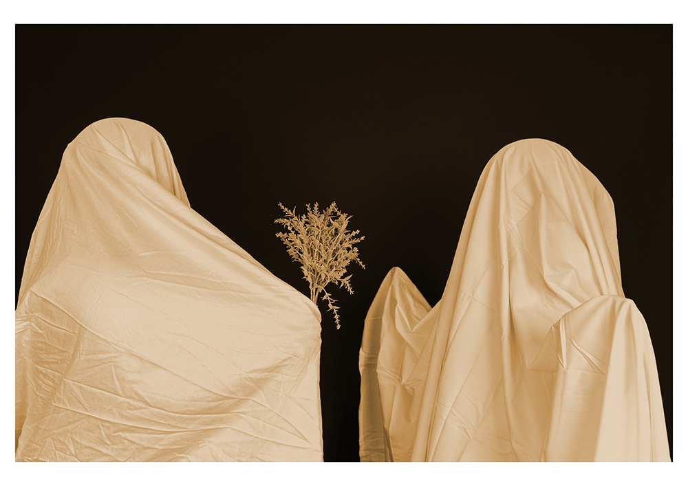 Two people in white sheets, one holding out flowers to the other