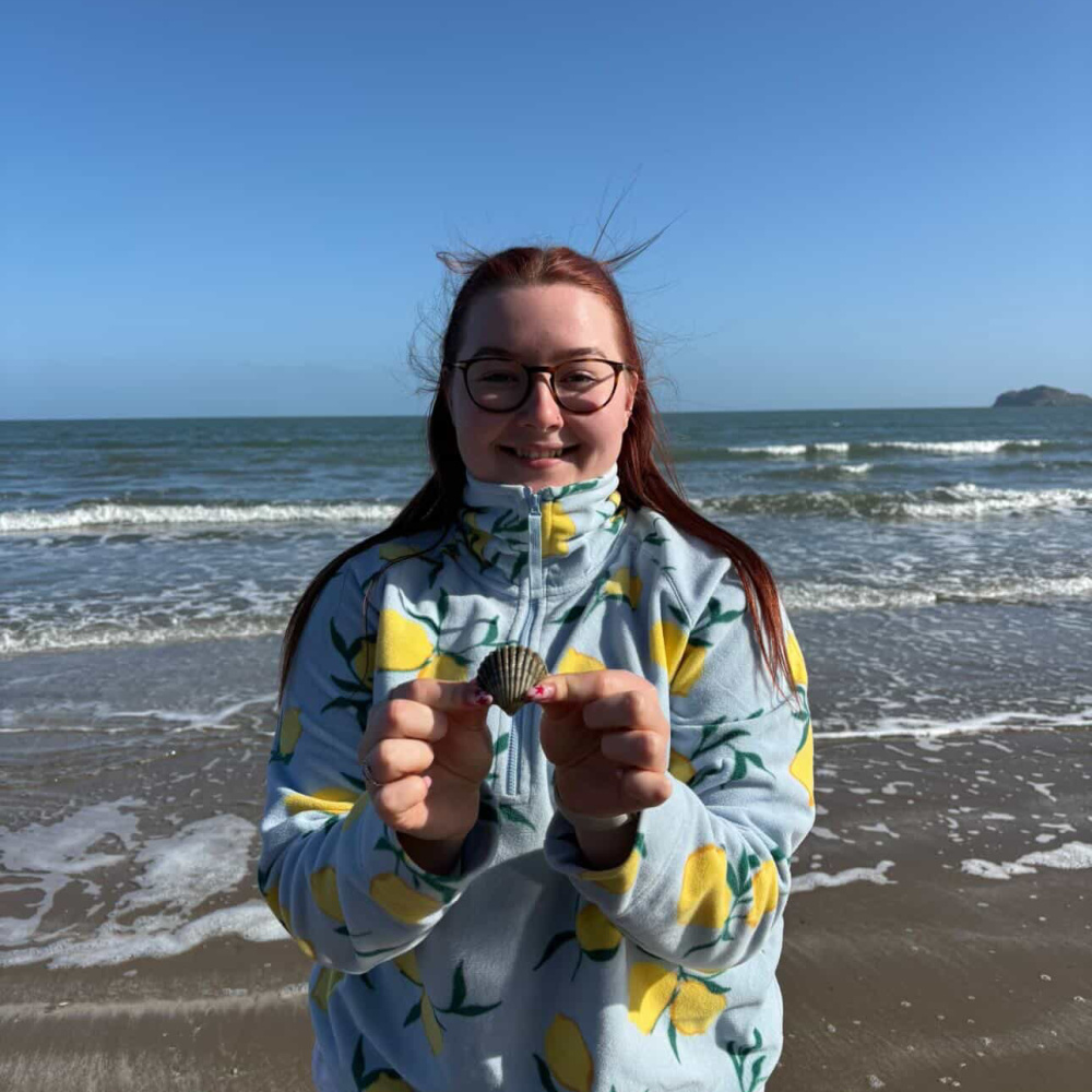 Smiling young woman with red hair and glasses standing on a beach, holding up a small scallop shell, wearing a light blue fleece with a lemon print pattern. Waves and a clear blue sky in the background.