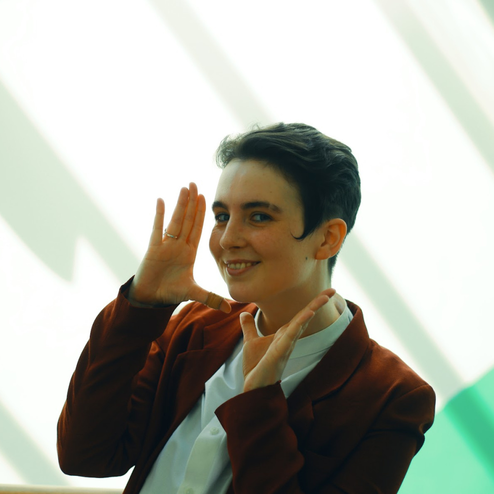A headshot of a nonbinary white person framing their head playfully with their hands while grinning. They have short brown hair with long sidelocks in curled points against their cheekbones. They are wearing a white shirt and orange blazer. 
